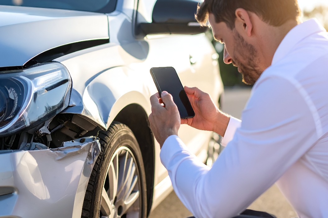 Ein Schadensregulierer fotografiert mit seinem Smartphone eine verbeulte Stoßstange; das Auto steht am Straßenrand, das Licht ist klar und nachmittäglich, der Fokus liegt auf dem Dokumentationsprozess